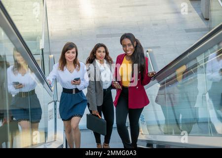 Tre giovani donne d'affari sulla scala mobile sul loro modo di lavorare con i loro dispositivi elettronici Foto Stock