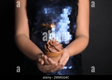 Donna in possesso di cupcake di compleanno con brillantante su sfondo nero, primo piano Foto Stock