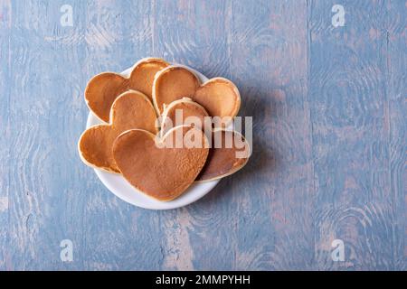 Frittella fatta in casa a forma di cuore su un piatto bianco su un tavolo di legno blu, spazio copia, vista dall'alto. Originale e creativa colazione di San Valentino Foto Stock