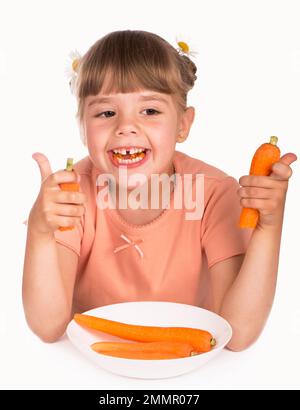 Problemi di odontoiatria pediatrica. La bambina in una t-shirt arancione e i fiori nei suoi capelli mangia una carota su uno sfondo bianco Foto Stock