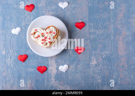 Colazione creativa per San Valentino. Pancake fatto in casa a forma di cuore, foderato con una pila e decorato con crema bianca su un piatto bianco su un legno blu Foto Stock