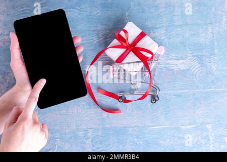 Una mano femminile tiene un telefono cellulare e un dito preme su uno schermo nero vuoto per lo shopping online e un carrello con una scatola regalo. Shopping online Foto Stock