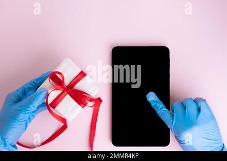 Una mano in un guanto blu protettivo preme su uno schermo nero vuoto di un tablet o telefono, un'altra mano in un guanto protettivo tiene un regalo su una schiena rosa Foto Stock