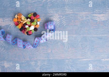 Concetto di controllo del peso. Concetto di alimento di dieta. Frutta secca e noci a forma di cuore e un metro a nastro assomiglia a un sorriso su una schiena di legno blu Foto Stock