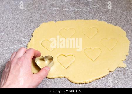 La mano di una donna taglia un biscotto a forma di cuore dalla pasta gialla su un tavolo da cucina grigio di tendenza, primo piano, vista dall'alto. Sorpresa per i cari a San Valentino Foto Stock