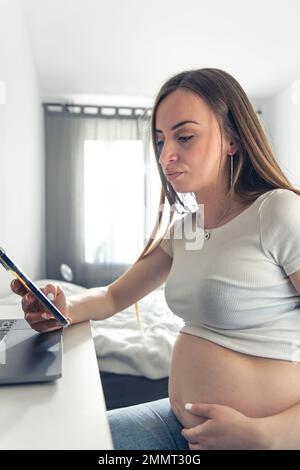 Una giovane donna incinta usa lo smartphone e il computer portatile a casa. Foto Stock