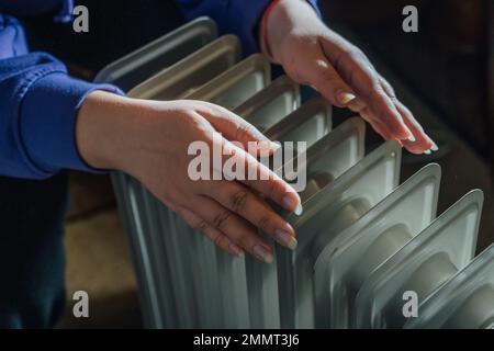 Le mani femminili si riscaldano sul riscaldatore dell'olio a casa da vicino. Inverno freddo con radiatore. Stagione di riscaldamento. Foto Stock