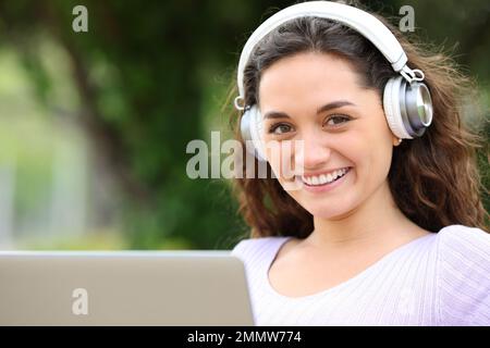 Una donna felice che usa il notebook e le cuffie ti guarda in un parco Foto Stock