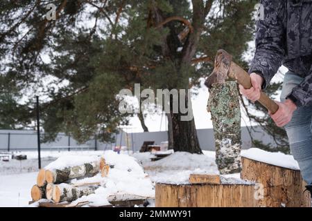 Un uomo trita legna da ardere con un'ascia in inverno all'aperto nella neve. Riscaldamento alternativo, raccolta del legno, crisi energetica Foto Stock