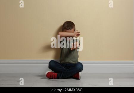 Piccolo ragazzo spaventato che chiude la faccia con la mano sul pavimento vicino al muro giallo. Bambini in pericolo Foto Stock