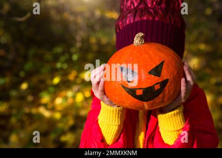 Una ragazza in caldo vestito rosso e giallo tiene in mano una zucca con gli occhi e la bocca dipinte. Halloween, Jack Lantern, senza volto. Umore autunnale, Foto Stock