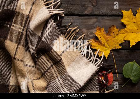 la plaid marrone di lana in una gabbia giace su un vecchio pavimento di legno come sfondo, uno sfondo di legno e su di esso una coperta e foglie autunnali, umore autunnale Foto Stock