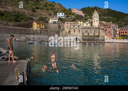 Italia, Liguria, Parco Nazionale delle cinque Terre Patrimonio Mondiale dell'UNESCO, villaggio di Vernazza, giovani che si tuffano nel porto e la chiesa parrocchiale di Santa Margherita d'Antiochia (Santa Margherita) fiancheggiata da un alto campanile ottagonale Foto Stock