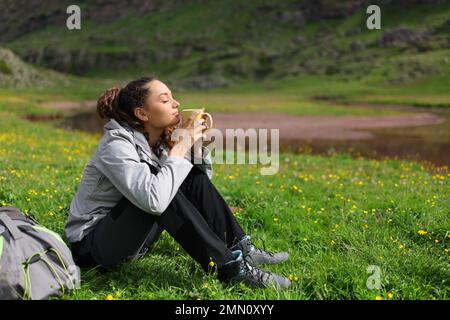 Escursionista godendo di una bevanda calda seduta sull'erba in natura Foto Stock