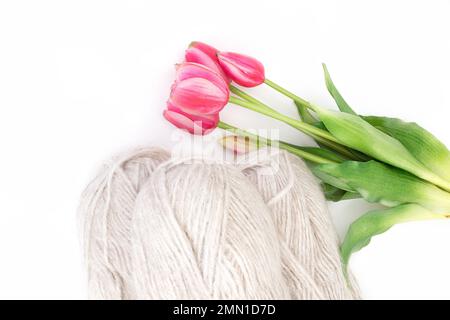 Gruppo di lana grigia di filato e filo con tulipano rosa su fondo bianco isolato. Maglieria, hobby, inverno. Spazio di copia. Vista dall'alto. Donne internazionali Foto Stock