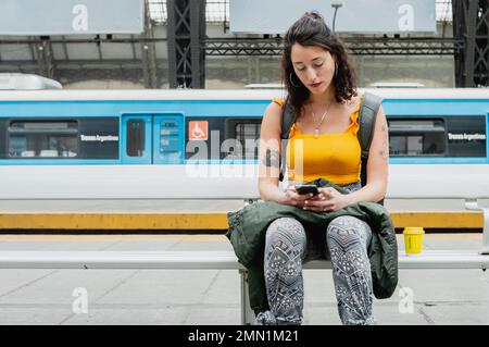 giovane donna latina di etnia argentina, seduta tranquilla alla stazione ferroviaria che controlla l'orario di imbarco sul telefono, concetto di viaggio, spazio copia. Foto Stock