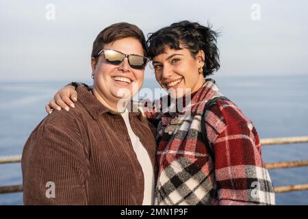 Due giovani donne, una brunetta e una persona non binaria curvy con un piercing al naso e capelli corti, sorridere e abbracciarsi mentre guardano nel venuto Foto Stock