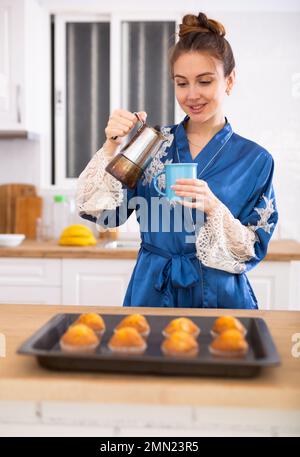 Donna in accappatoio blu che versa caffè vicino al tavolo con muffin al forno Foto Stock