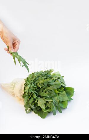 Rucola verde fresca e spinaci in un sacchetto a rete ecologico su superficie bianca. La mano della donna tiene una foglia di rucola. La vista è diretta. Concetto di alimentazione sana. Scheda con spazio di copia per il testo. Foto Stock