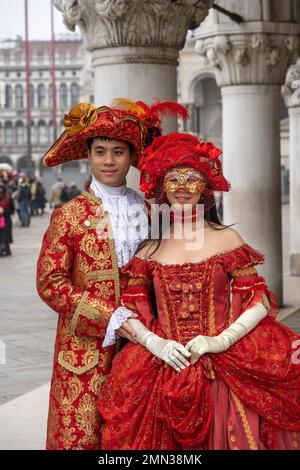Giovane asiatico e giovane donna in costume di carnevale rosso medievale, cappelli con piume e maschere in Piazza San Marco a Venezia Foto Stock