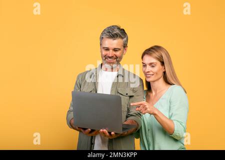 Eccitata coppia caucasica di mezza età che utilizza un notebook insieme, donna che guarda e indica il gadget, sfondo giallo Foto Stock