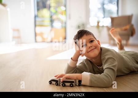 Allegro pensive bambino europeo in pigiama giace sul pavimento, gioca con le auto da solo, godere di riposo e tempo libero Foto Stock