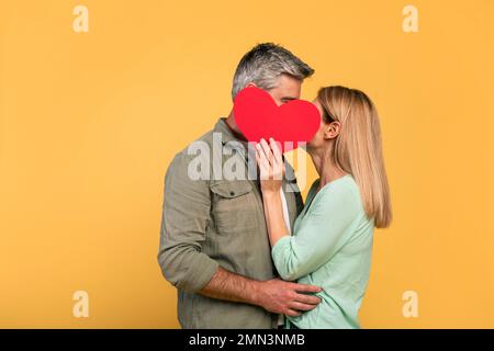 Irriconoscibile coppia di mezza età che si nasconde dietro un grande cuore rosso, baciandosi e abbracciando, sfondo giallo Foto Stock