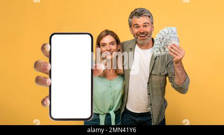 Concetto di omaggio. Coppia emotiva di mezza età che tiene un sacco di dollari in contanti, mostrando smartphone con schermo vuoto, mockup Foto Stock