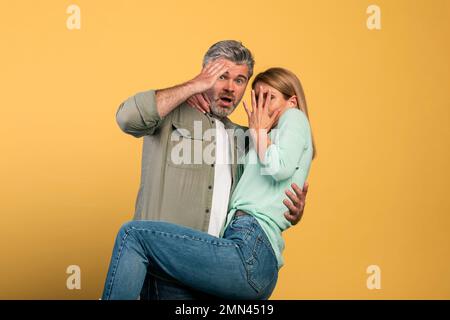 Ritratto di coppia di mezza età spaventata che copre la bocca e gli occhi, donna che salta sull'uomo, guardando la macchina fotografica Foto Stock