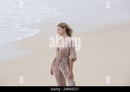 Ritratto di bella giovane donna in seta beige vestito su spiaggia di sabbia vuota, idilliaca vacanza estiva lontano dalla città Foto Stock