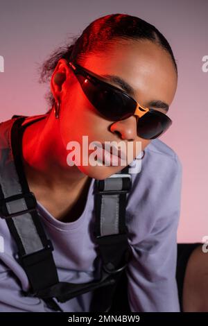 donna afroamericana in felpa e occhiali da sole eleganti in grigio con luce rossa Foto Stock