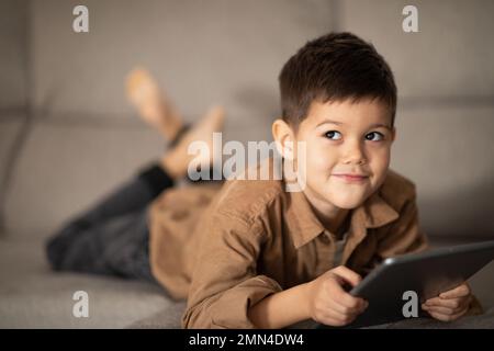 Piccolo ragazzo europeo, pensivo e allegro, si trova sul divano, pensa con il tablet, guarda lo spazio libero all'interno della stanza Foto Stock