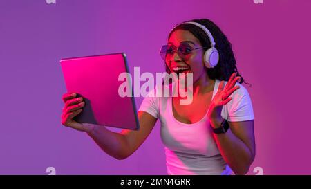 App straordinaria. Donna nera entusiasta nelle cuffie wireless che utilizzano il tablet digitale Foto Stock