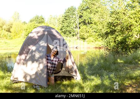 Una donna felice in una camicia a quadri guarda fuori da una tenda turistica in un'escursione sulla riva del fiume al mattino. Campeggio in natura, pernottamento in famiglia Foto Stock