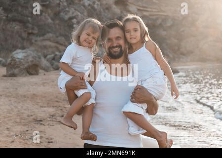 Amore e cura di papà. Il padre tiene in braccio due figlie, forza e coraggio. Famiglia amichevole a casa e in vacanza. Foto Stock