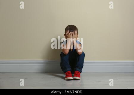 Piccolo ragazzo che chiude il viso con le mani sul pavimento vicino al muro giallo. Bambini in pericolo Foto Stock