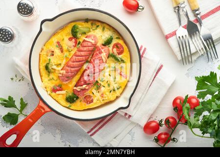 Omelette o frittata con due salsicce, formaggio fuso, pomodori ciliegini, broccoli e prezzemolo su padella rossa o teglia di ferro su fondo bianco di cemento vecchio Foto Stock