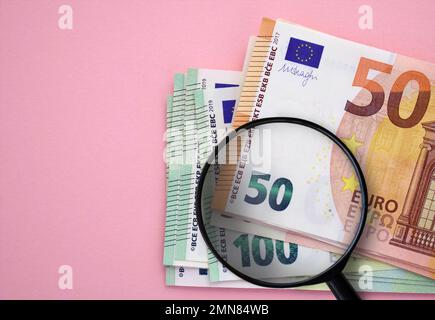 Confezione di banconote in euro (50 euro, 100 euro) e lente di ingrandimento su sfondo rosa. Valuta europea, risparmio, ricchezza Foto Stock