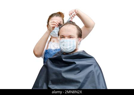 Una donna in maschera medica taglia i capelli di un uomo con forbici, isolato su uno sfondo bianco Foto Stock