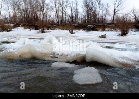 Il primo debole ghiaccio autunnale sorge in onde da una forte corrente e vento d'acqua vicino alla riva con alberi. Foto Stock
