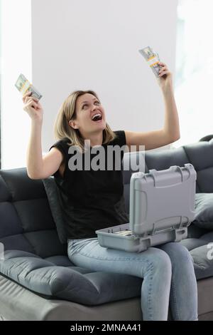 Donna felice che tiene pile di banconote in dollari in mano mentre si siede sul divano con valigia piena di soldi Foto Stock