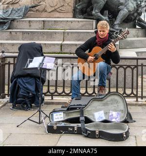 Un uomo biondo di mezza età che indossa un cappotto nero corto e jeans blu suona una chitarra seduto in strada con un supporto musicale e una borsa da concerto aperta davanti a lui Foto Stock