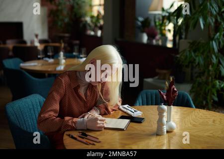 Giovane donna albina in casualwear intelligente schizzo in blocco note con matita mentre si siede al tavolo in un accogliente bar o ristorante e in attesa di un amico Foto Stock