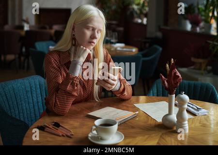 Ragazza albina riposante in elegante casualwear guardando lo schermo dello smartphone mentre si SMS o scorrere i dati online mentre si è seduti in un bar Foto Stock