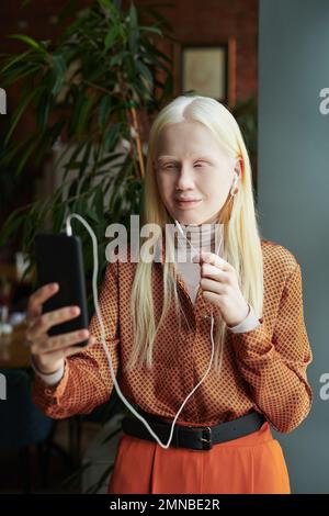 Giovane ragazza albina in blusa intelligente che parla con un amico in videochat mentre si trova davanti alla fotocamera contro la pianta verde nel bar Foto Stock