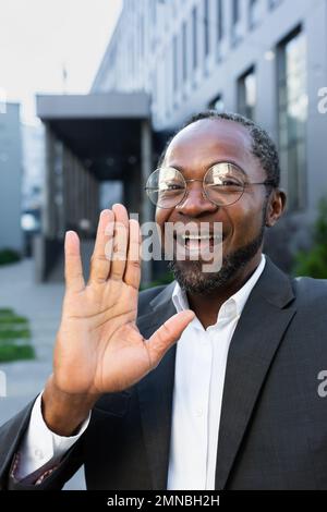 Primo piano. Uomo afroamericano sorridente in tuta e occhiali in piedi vicino al centro ufficio che parla durante una videochiamata. Si fa una mano alla telecamera, salutando. Foto Stock