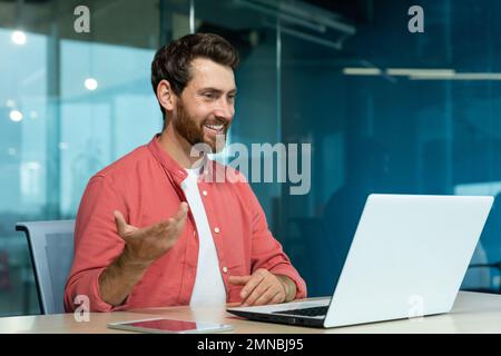 Imparare online. Un giovane insegnante maschile in una camicia rossa siede in ufficio al tavolo, lavora su un computer portatile, insegna attraverso una videochiamata, conferenza, webinar. Spiega, sorride. Foto Stock