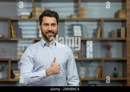 Primo piano. Ritratto di un giovane uomo d'affari di successo, avvocato. In piedi in una camicia bianca in ufficio, guardando la macchina fotografica, puntando super, sorridente. Foto Stock