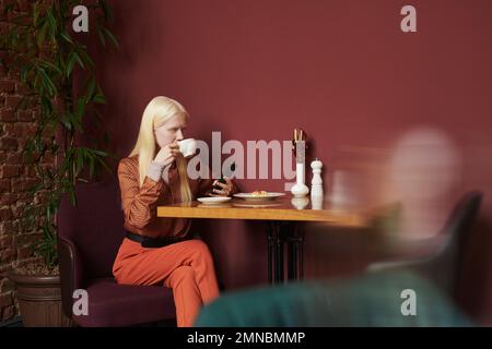 Ragazza albina riposante in elegante casualwear con caffè o tè con dessert e scorrimento in gadget mobile mentre si siede al tavolo in accogliente caffè Foto Stock