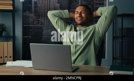 Felice uomo latino-americano finito il lavoro sul portatile soddisfatto giovane uomo d'affari riposo seduto in ufficio con le mani dietro la testa sensazione alleviato dopo il lavoro Foto Stock